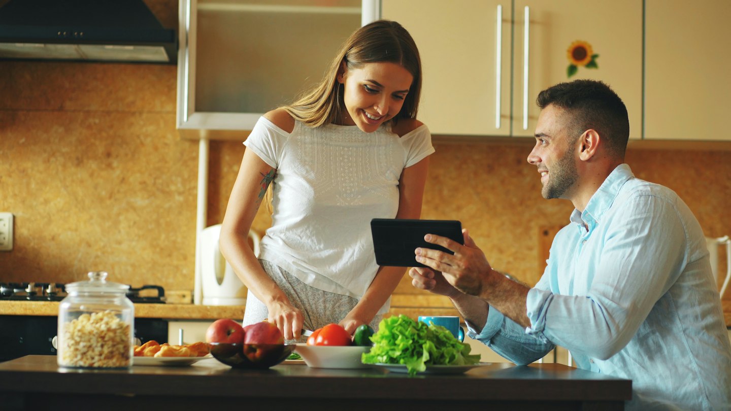 A man shows a digital tablet to a woman preparing vegetables.