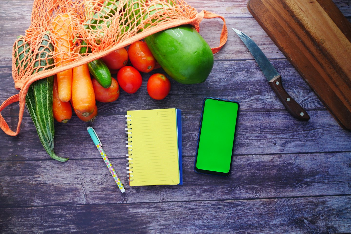 Fresh produce spills from an orange mesh bag next to a notebook and phone.